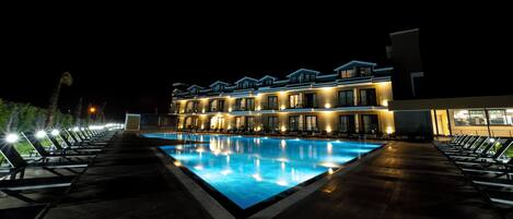 Una piscina grande y rectangular con cubierta de madera y sillones de descanso a ambos lados, rodeada por un edificio de varios pisos con balcones y ventanas iluminadas.