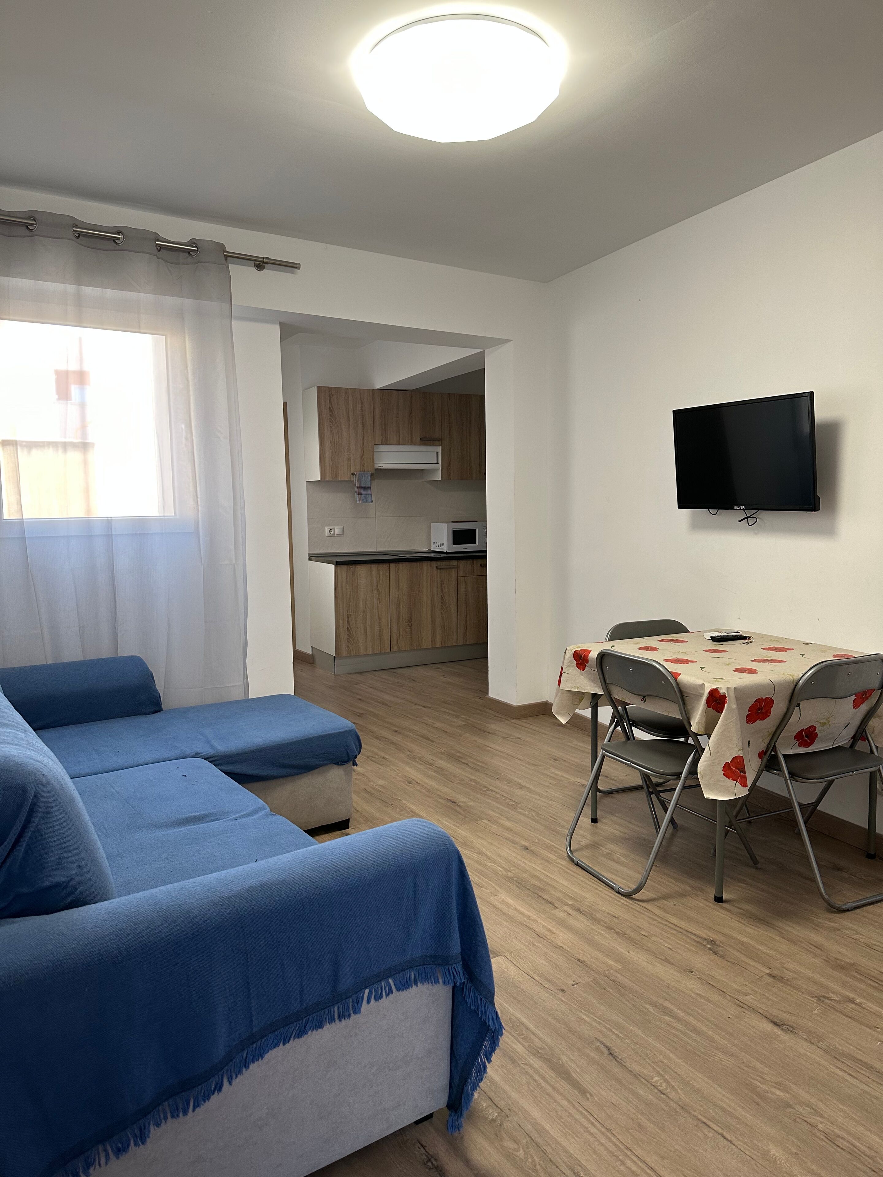 A modern living room with a sofa, a dining table with chairs, a flat-screen TV, and a kitchen in the background.