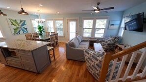 A spacious kitchen with a granite island, wooden dining table, and chairs. There is a gray sofa and patterned armchair in the living area. - M931- Sound Side, Corolla, Saturday Check In, Comm. Pool! (Corolla)