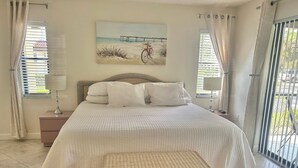 A bedroom with a bed, two nightstands, a wicker side table, and a beach-themed painting on the wall. - Serene retreat awaits at prime location in charming Timberwoods/Village des Pins (Sarasota)