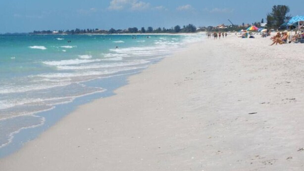 A beach with clear turquoise water, a sandy shore, and umbrellas and people in the distance. - Serene retreat awaits at prime location in charming Timberwoods/Village des Pins (Sarasota)