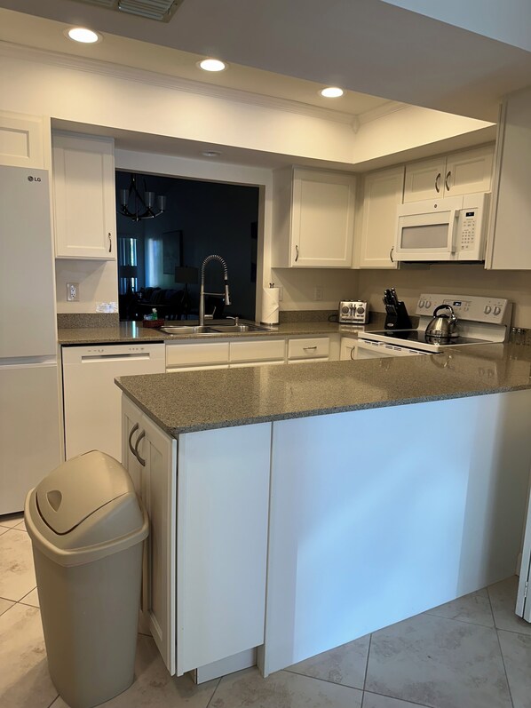 A modern kitchen with white cabinetry, a granite countertop, a stainless steel sink, a microwave, a kettle, and a dishwasher. - Serene retreat awaits at prime location in charming Timberwoods/Village des Pins (Sarasota)