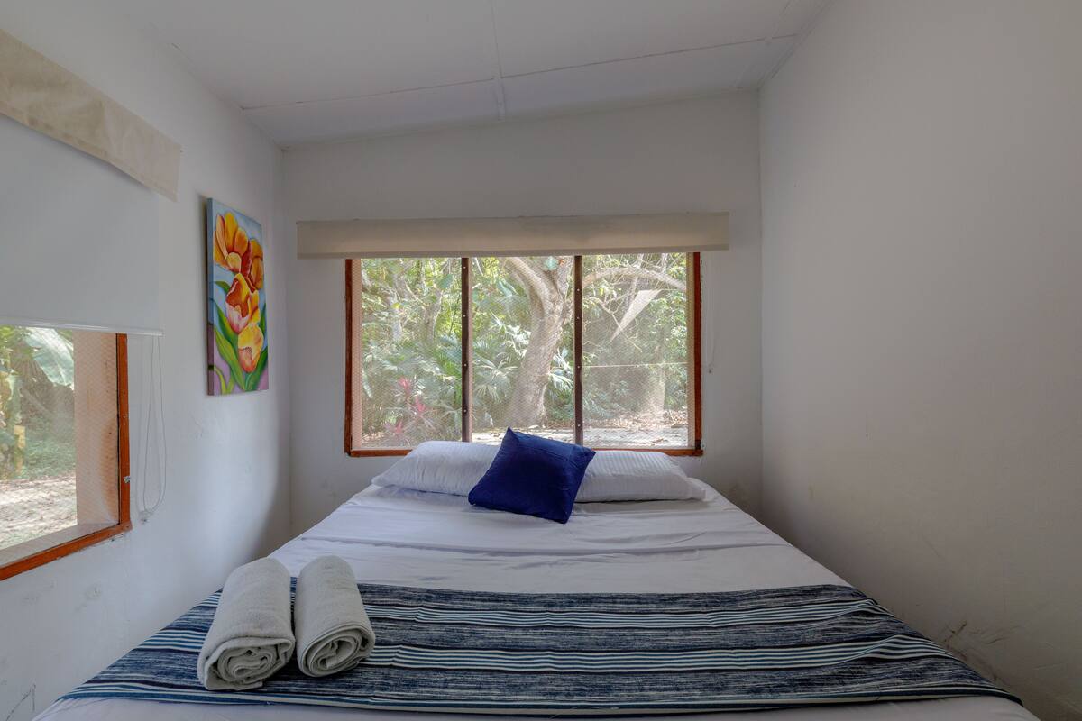 A bedroom with a bed, a window with a view of trees, and a painting on the wall.