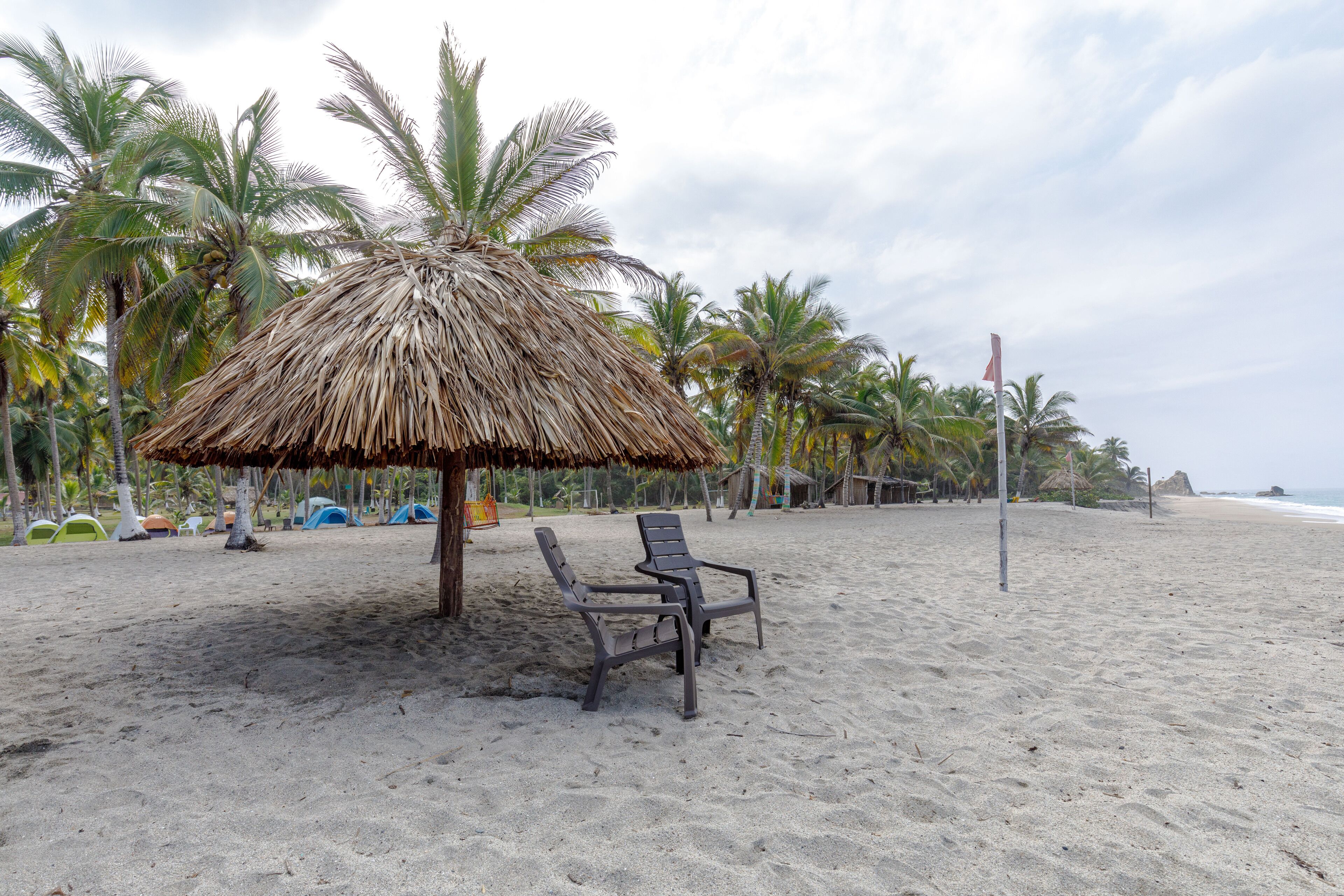 A beach with a thatched umbrella, two lounge chairs, and palm trees.