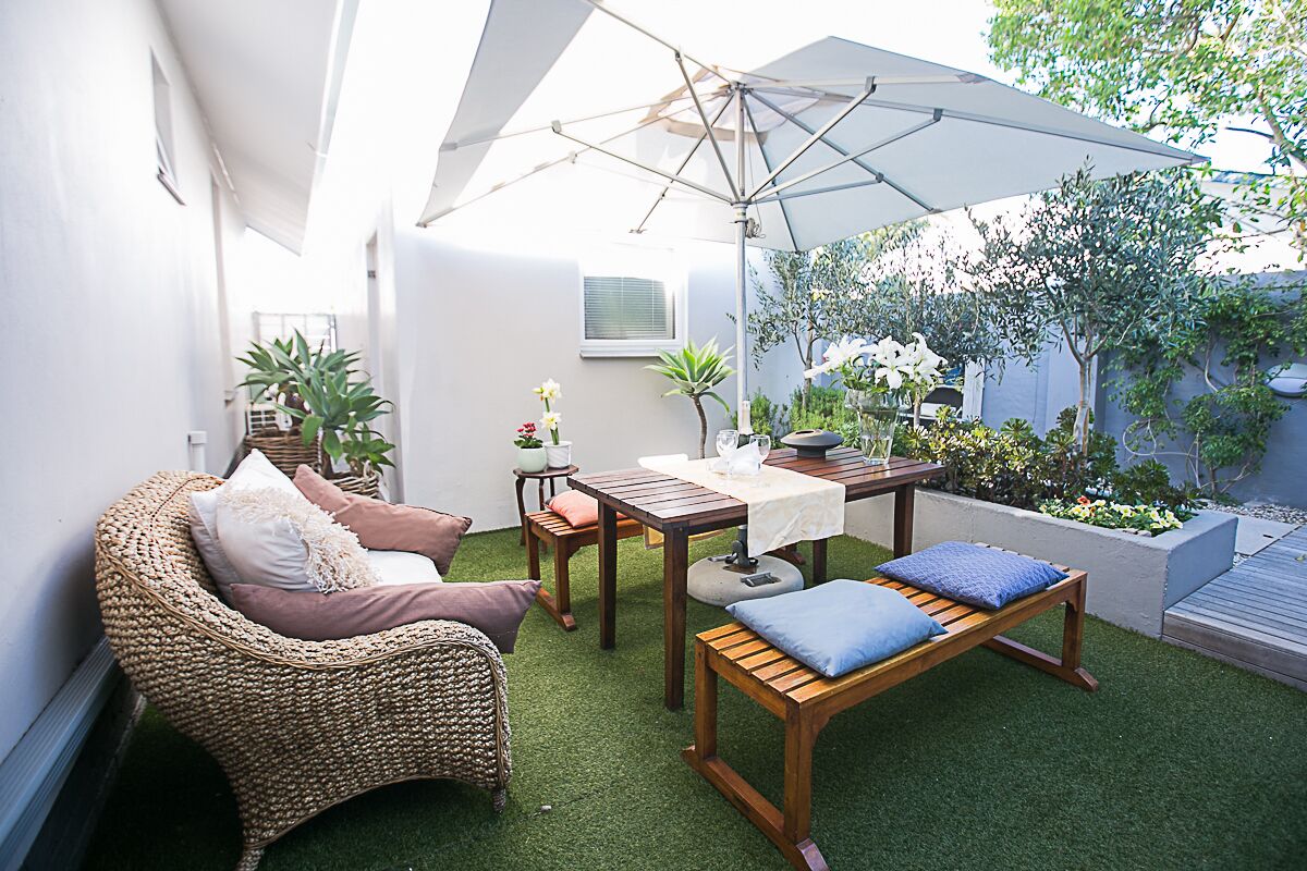 A cozy outdoor seating area with a wicker chair, wooden bench, and table, surrounded by greenery and a white umbrella.