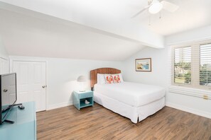 Une chambre à coucher comprenant un lit, une table de chevet, un bureau, un ventilateur de plafond et une fenêtre avec des stores.