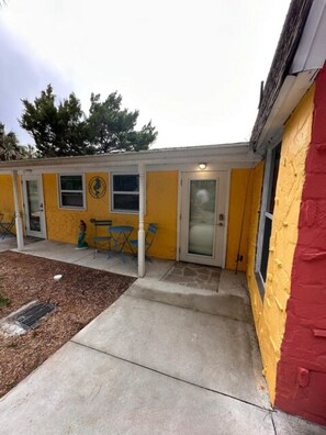 Un bâtiment jaune avec une partie rouge, une porte et un petit patio agrémenté de chaises bleues.