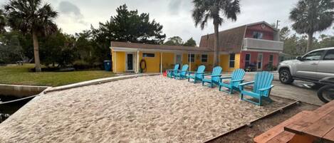 A sandy beach area with blue chairs and a picnic table, surrounded by buildings and palm trees.
