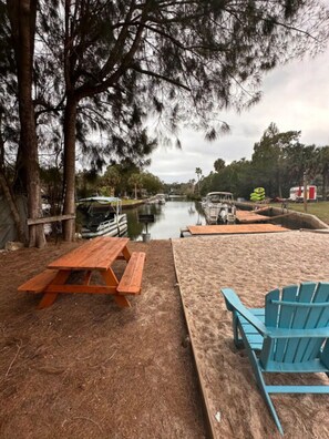 Un espace de pique-nique au bord de la rivière, avec des bancs en bois et une chaise Adirondack bleue, entouré d’arbres et bordé d’un cours d’eau paisible.