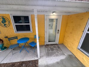 Un mur extérieur jaune avec une porte blanche et une fenêtre. On y voit des chaises pliantes bleues et une petite table à l’extérieur.