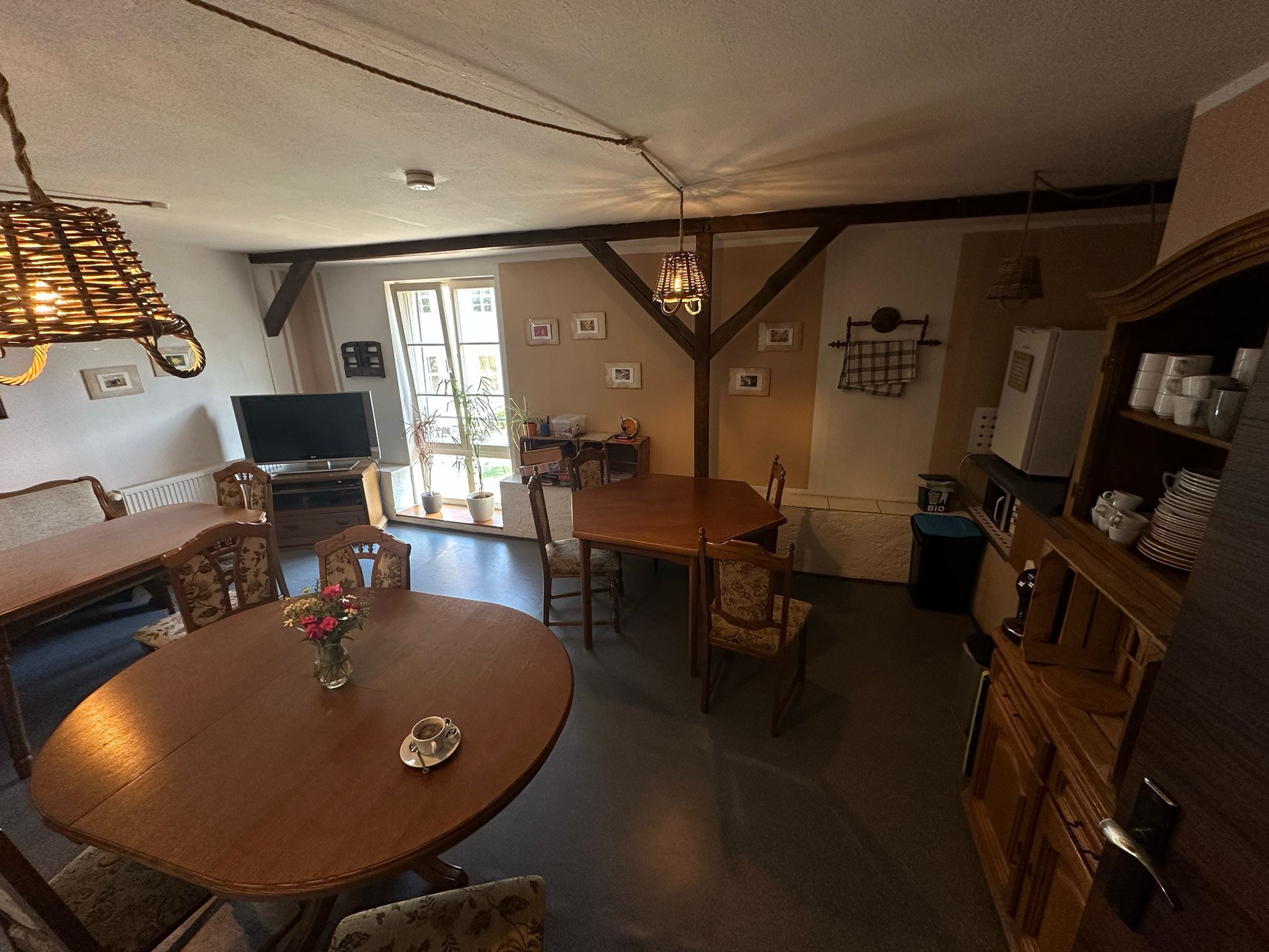 A dining area with a round wooden table and chairs, a television in the background, and a kitchen area with cabinets and a refrigerator.