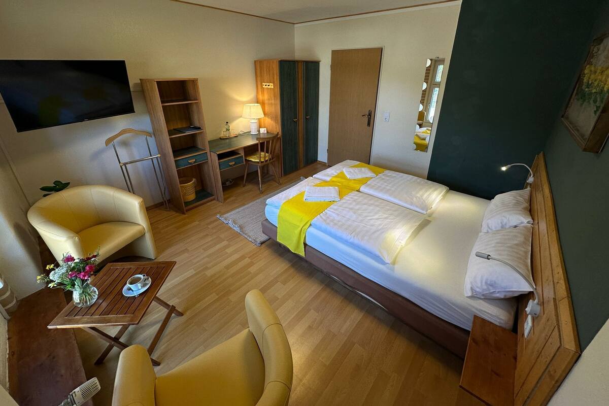 Quarto de hotel com cama, escrivaninha, cadeira, televisão e uma pequena mesa com um vaso de flores.