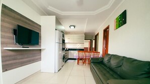 A modern living room with a sofa, a television mounted on a wooden panel, a kitchen area with cabinets and a refrigerator, and a dining table with chairs. - Residencial Falcão (Bombinhas)