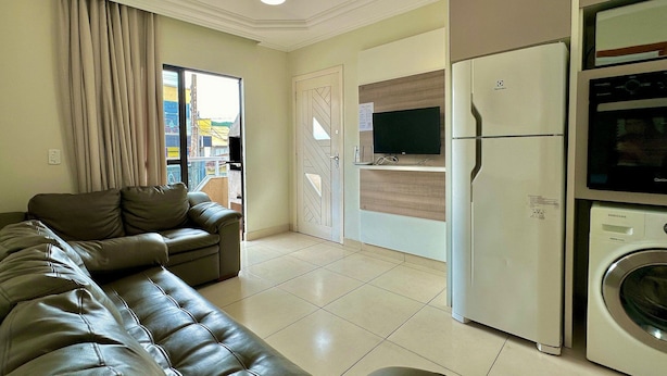 A living room with a leather sofa, a television, a refrigerator, and a washing machine. - Residencial Falcão (Bombinhas)