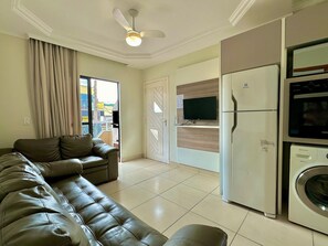 A living room with a leather sofa, a television, a refrigerator, and a washing machine. - Residencial Falcão (Bombinhas)