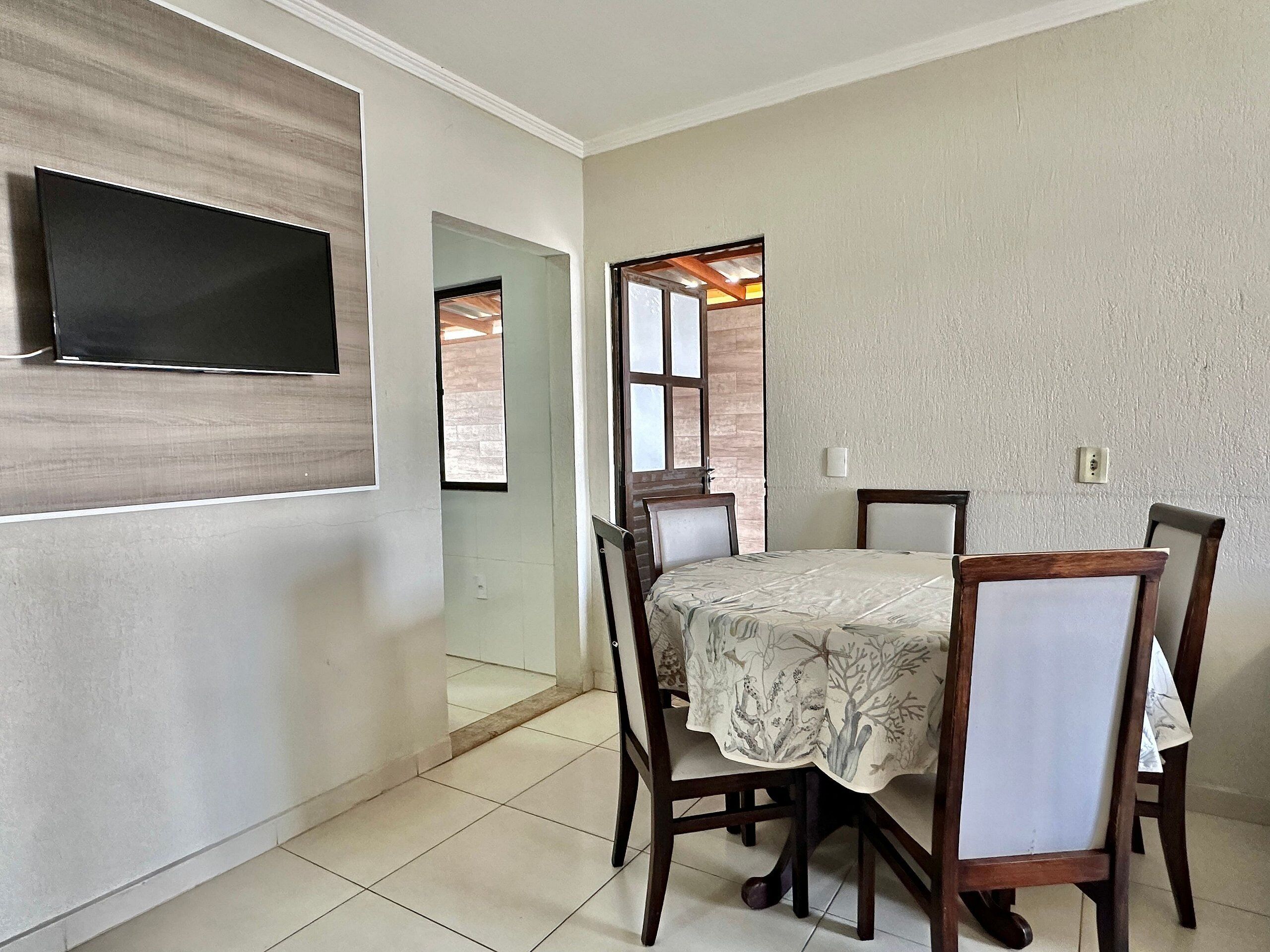 A dining area with a table and chairs, a flat-screen TV mounted on the wall, and a window with a view of the outside.