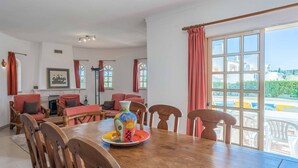 A dining area with a wooden table, chairs, and a colorful vase. There are windows with curtains, a fireplace, and a view of the outside. - Villa Noras - Three Bedroom Villa, Sleeps 6 (Albufeira)