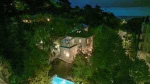 A large, modern house surrounded by dense greenery, with a swimming pool in the foreground.