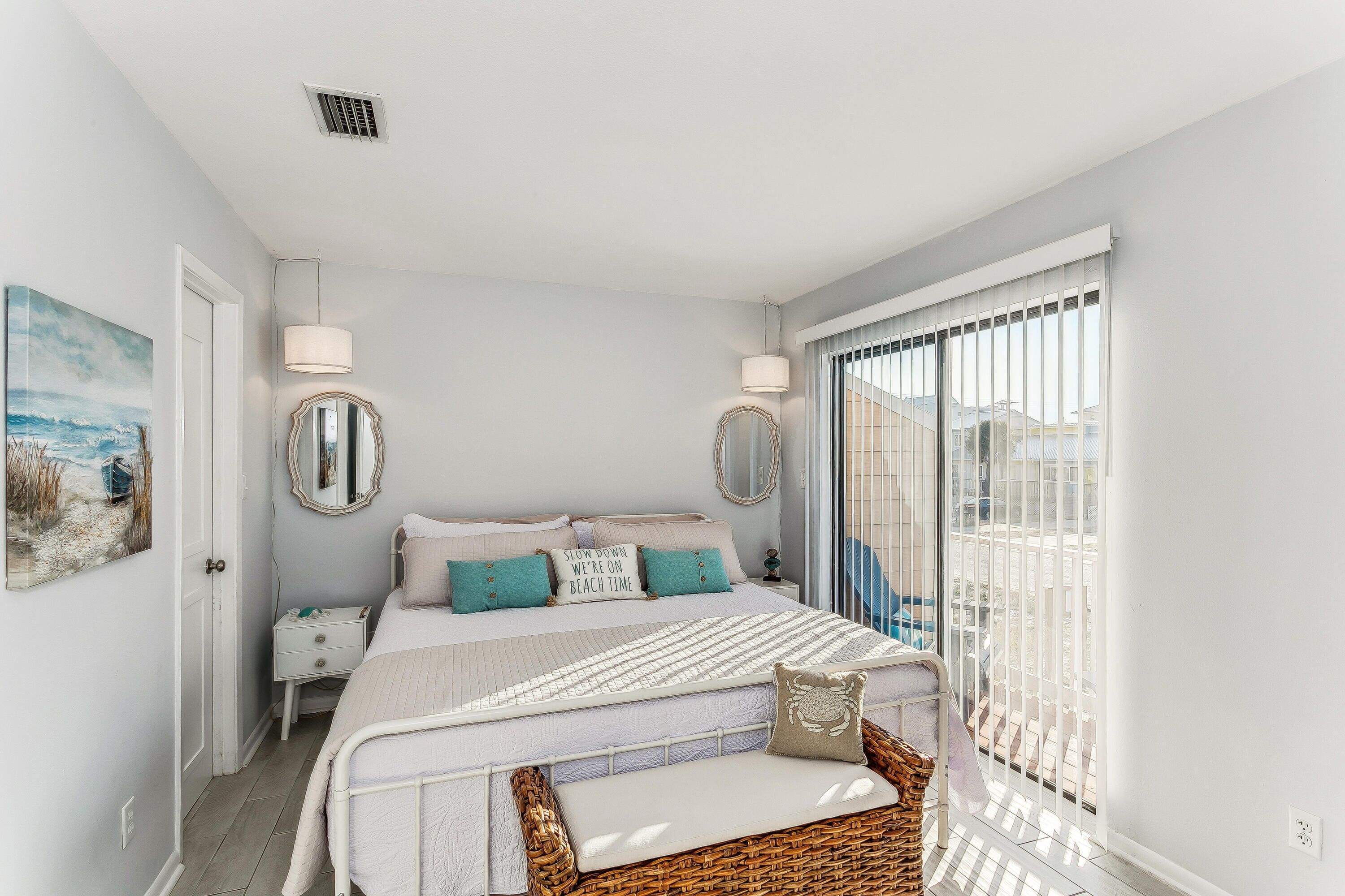 A bedroom with a bed, bedside tables, a mirror, a window with blinds, and a view of the outside.