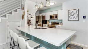 Une cuisine moderne dotée d’un grand îlot central, de tabourets de bar, d’appareils électroménagers en acier inoxydable et de luminaires suspendus.
