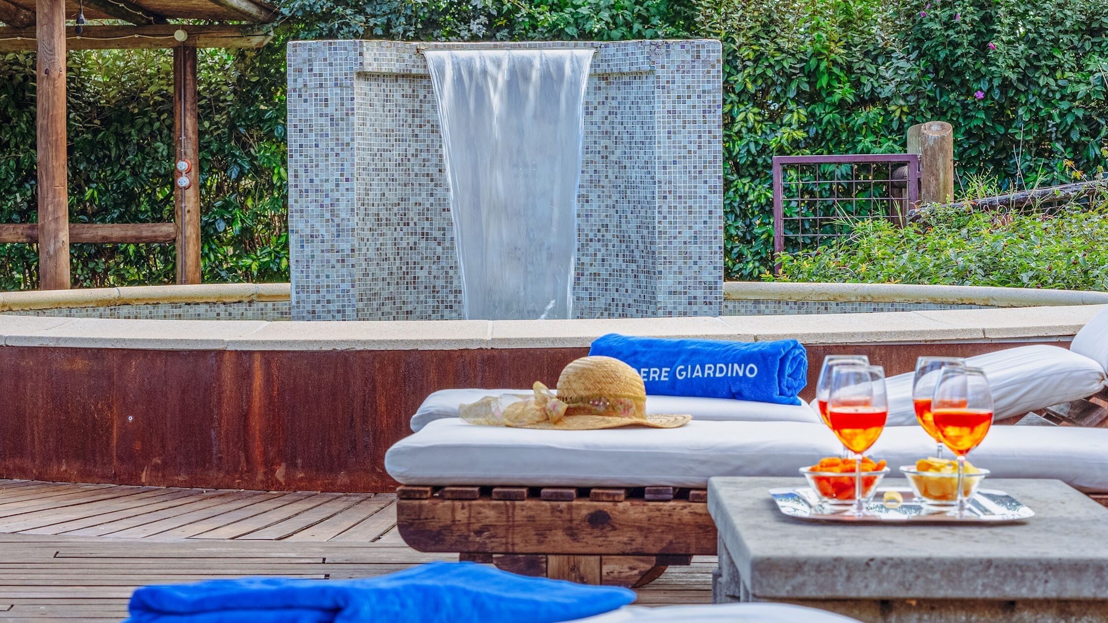 A poolside area with a mosaic wall, a wooden deck, lounge chairs, and a table with drinks and snacks.