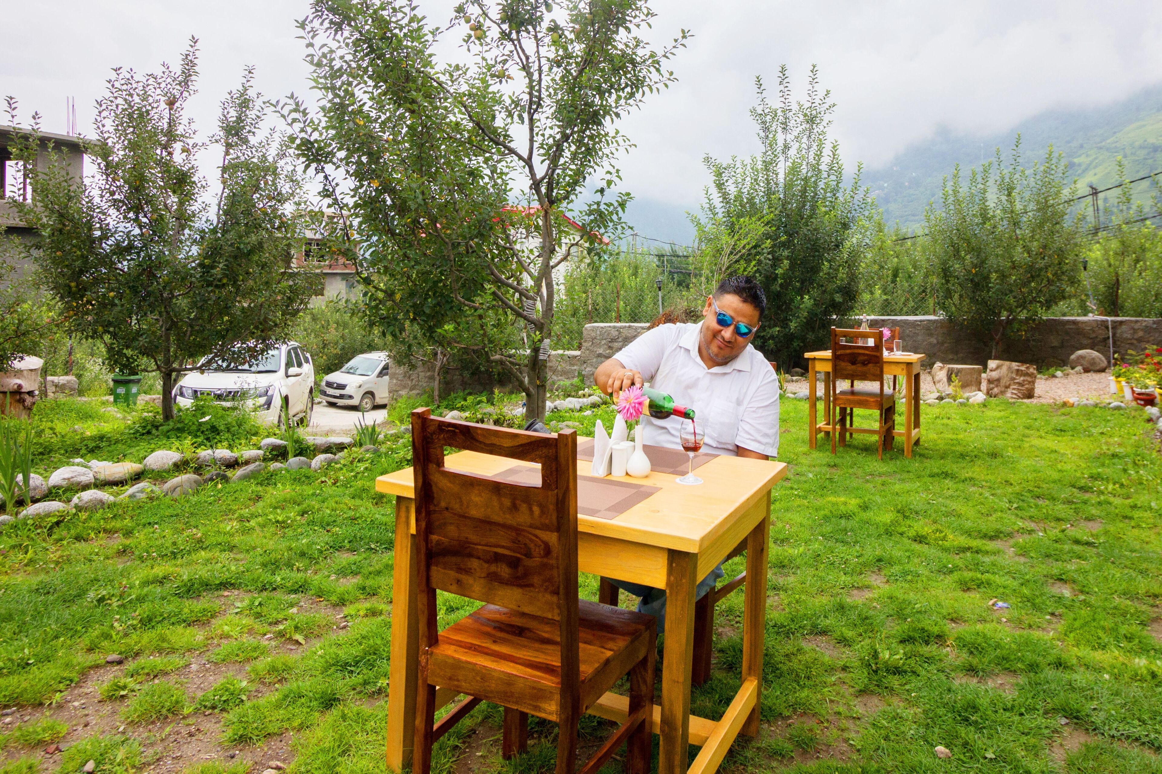 A person sitting at an outdoor wooden table with a bottle and glasses, surrounded by greenery and a stone wall.