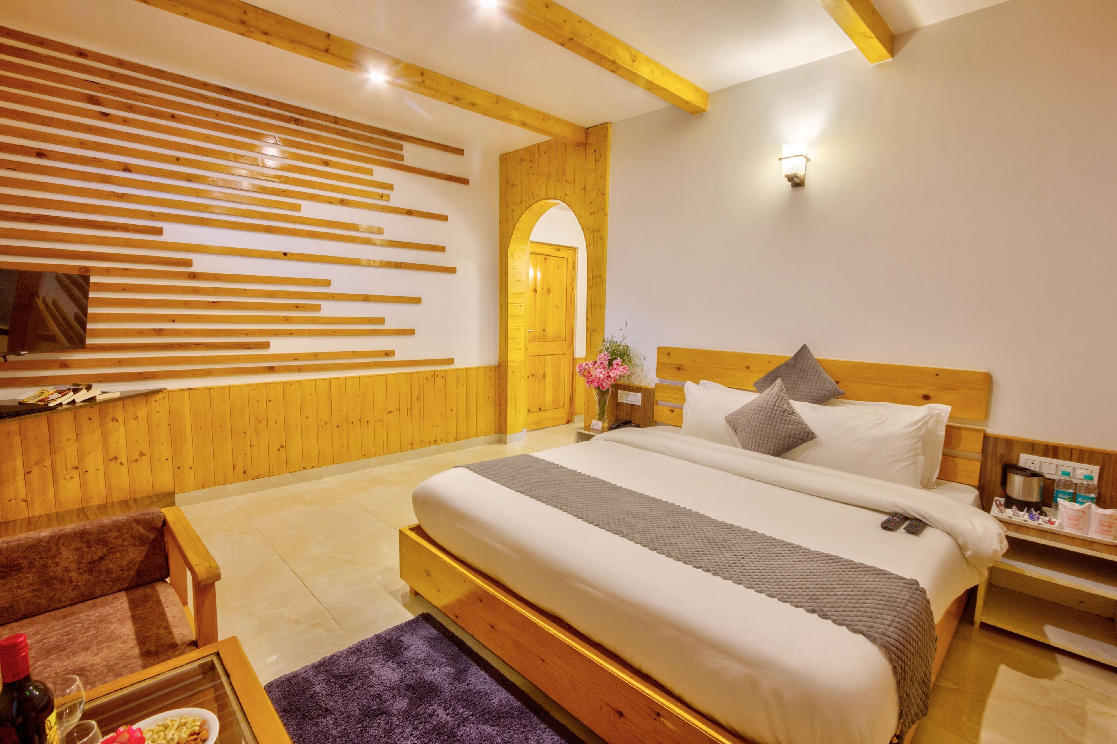 Une chambre d’hôtel moderne avec un mur en lambris de bois, un grand lit avec des oreillers, une table de chevet avec une machine à café et une petite table avec une bouteille et des verres.