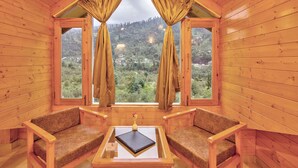 A wooden cabin room with two wooden chairs, a small table with a book and a vase, and large windows offering a view of a forested hillside. - La Aero Resort (Manali)