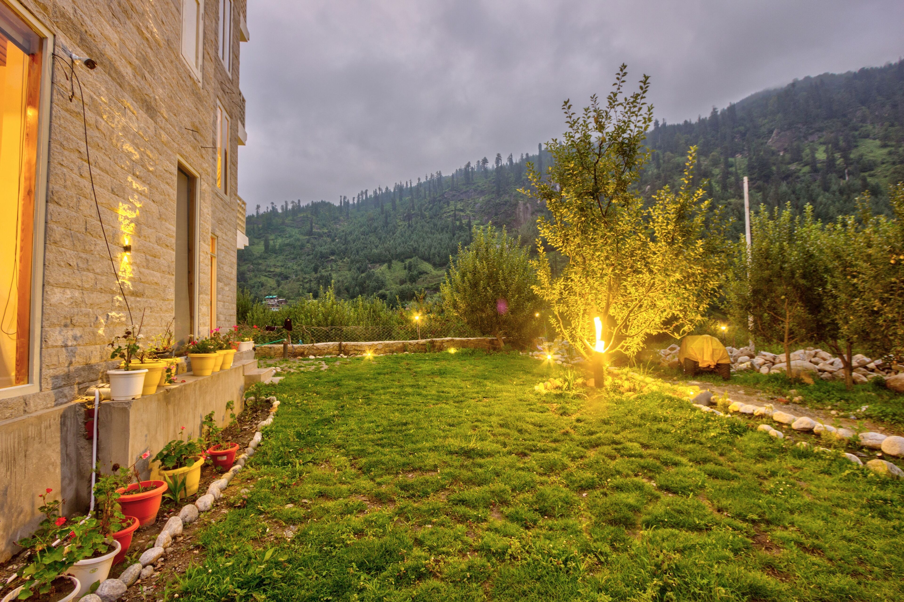 A stone building with a garden in front, potted plants, and a view of a forested hillside.