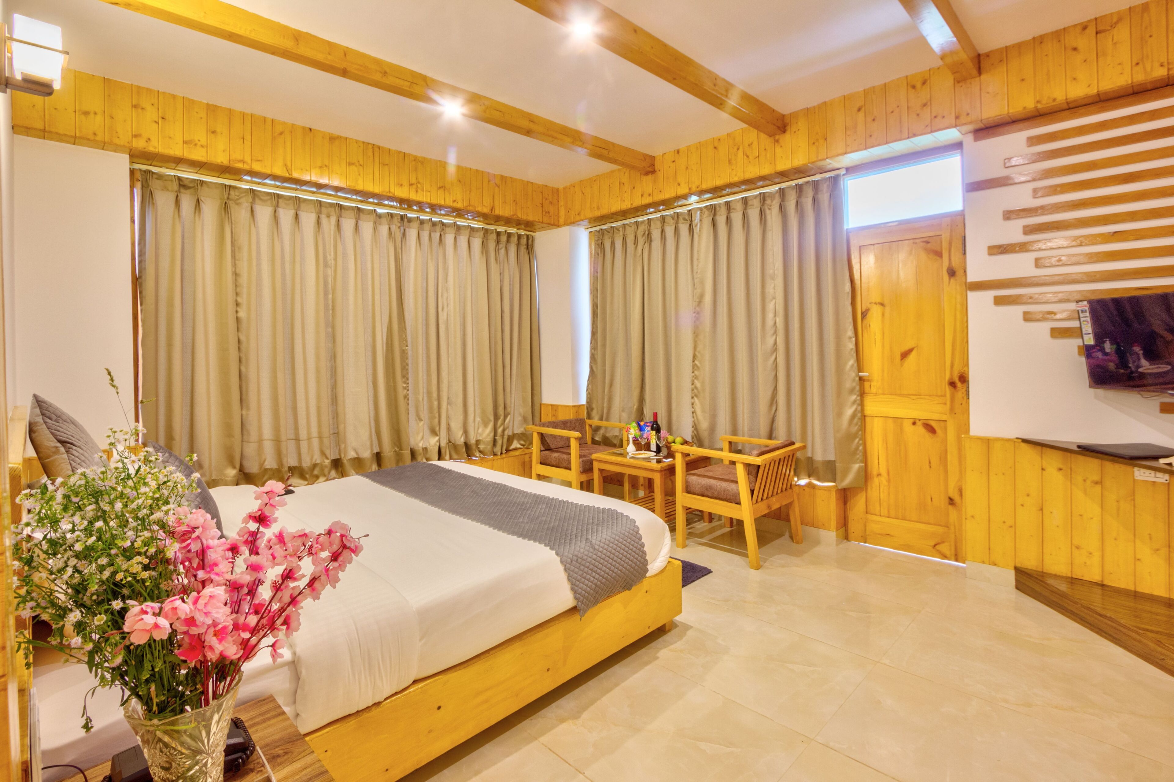 A bedroom with a wooden bed, a desk, and a chair. There is a window with curtains and a wooden door.