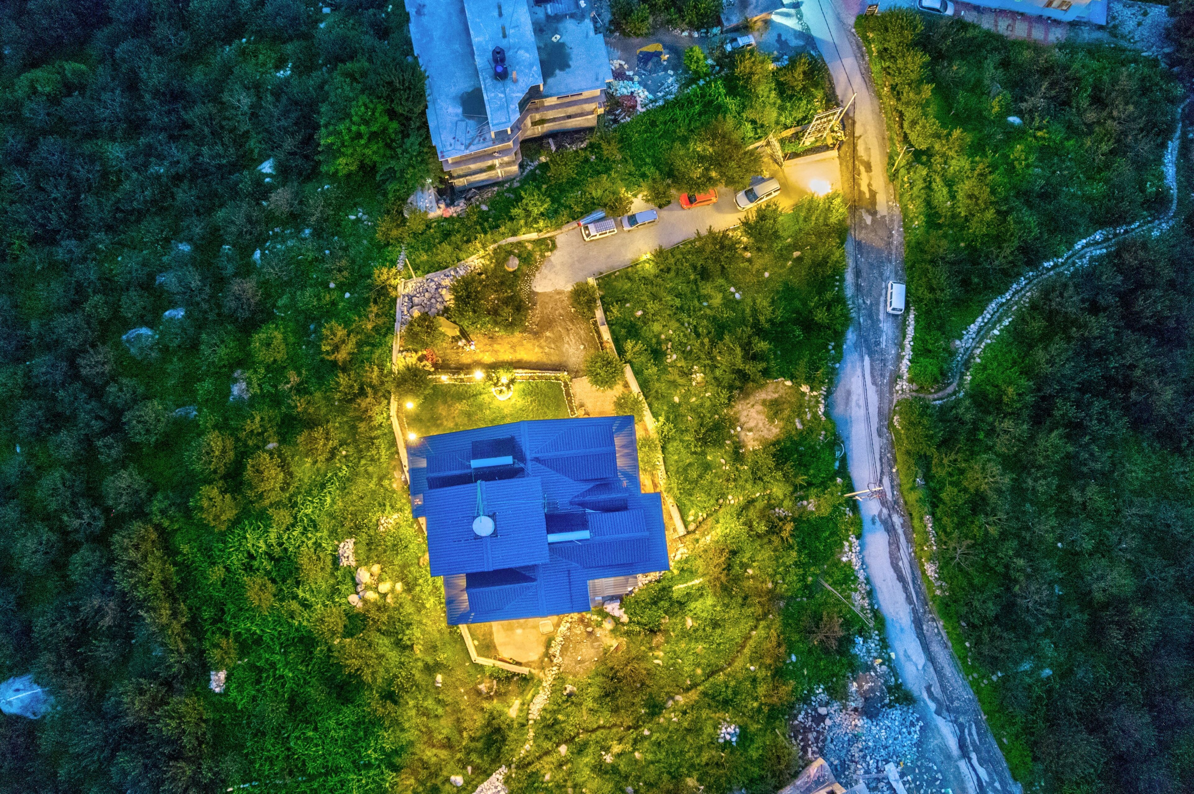 An aerial view of a building surrounded by dense greenery, with a parking area and a road nearby.