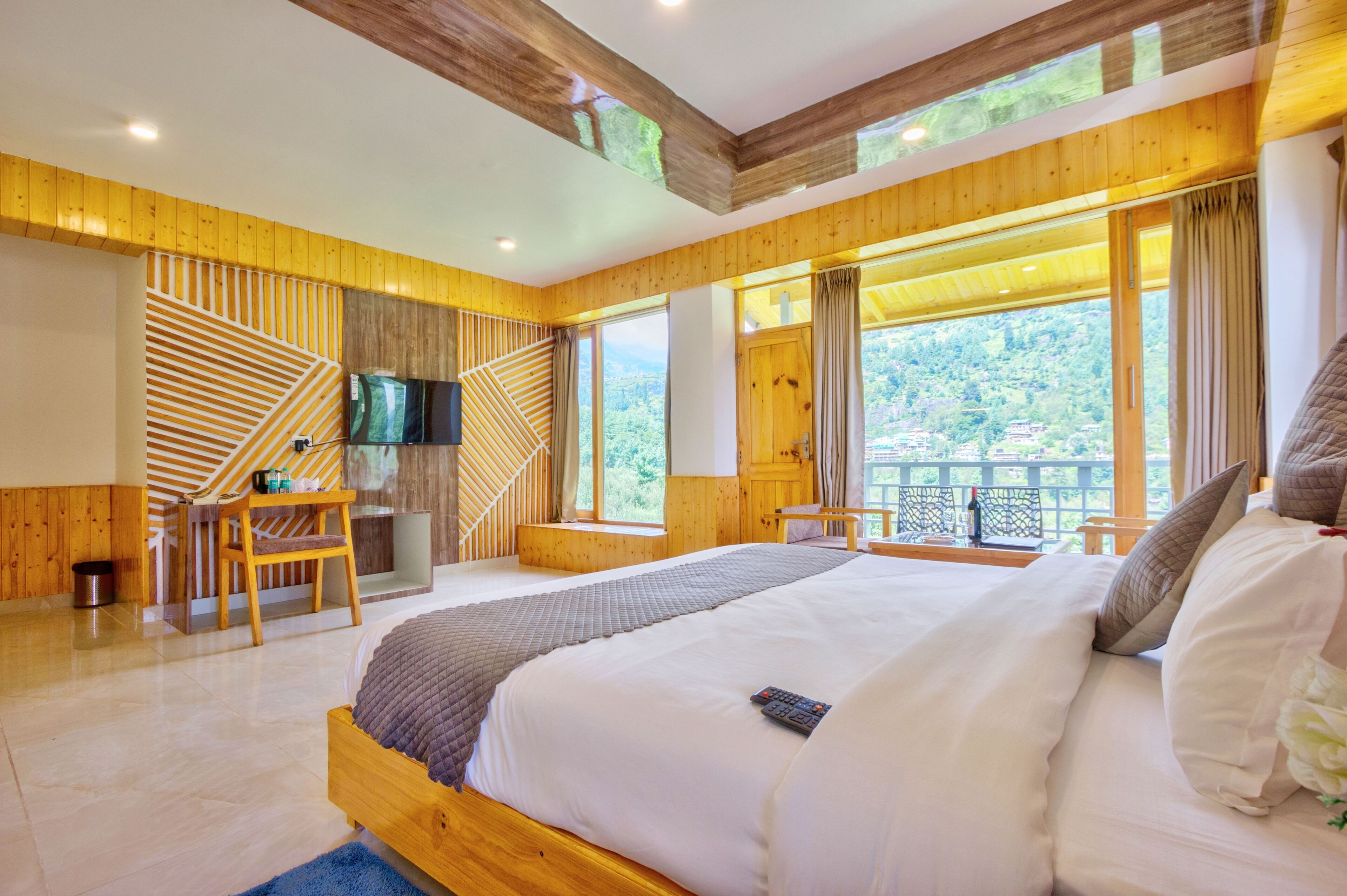 A spacious bedroom with a large bed, wooden walls, a TV, and a balcony with a view of the forest.