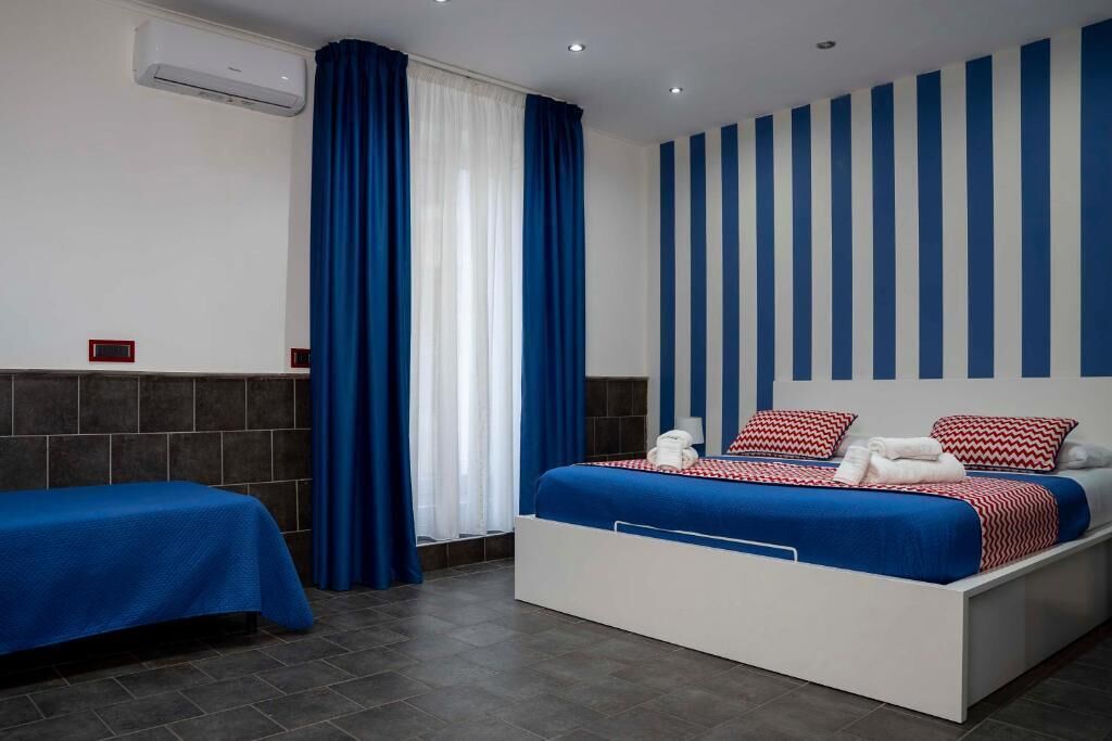 A hotel room with a bed, a bedside table, a window with curtains, a wall with vertical stripes, and an air conditioning unit.