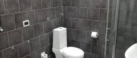 Un baño con azulejos oscuros, un inodoro, un bidé, un lavabo y una cabina de ducha con paredes de vidrio.