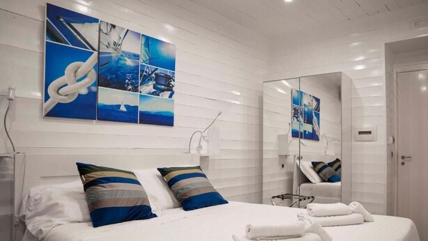 A hotel room with a large bed, white walls, and a mirror. There are multiple pillows on the bed and a small table with a lamp. - Yacht Club Angioino (Naples)
