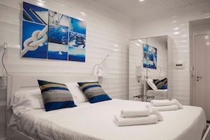 A hotel room with a large bed, white walls, and a mirror. There are multiple pillows on the bed and a small table with a lamp. - Yacht Club Angioino (Naples)