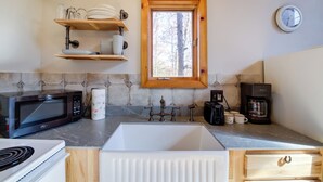 Una cocina con un lavabo blanco, un microondas negro, una cafetera y estantes con vajilla y cristalería.