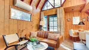A cozy cabin interior with a leather sofa, a wooden chair, a dining table, and a large window with curtains. - 3 Mi to Big Bear Village: Charming Sugarloaf Cabin (Sugarloaf)