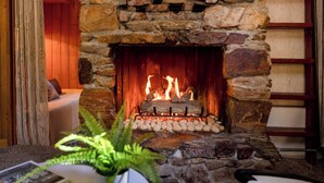 A stone fireplace with a fire, a white chair, and a fern plant in a cozy room. - 3 Mi to Big Bear Village: Charming Sugarloaf Cabin (Sugarloaf)