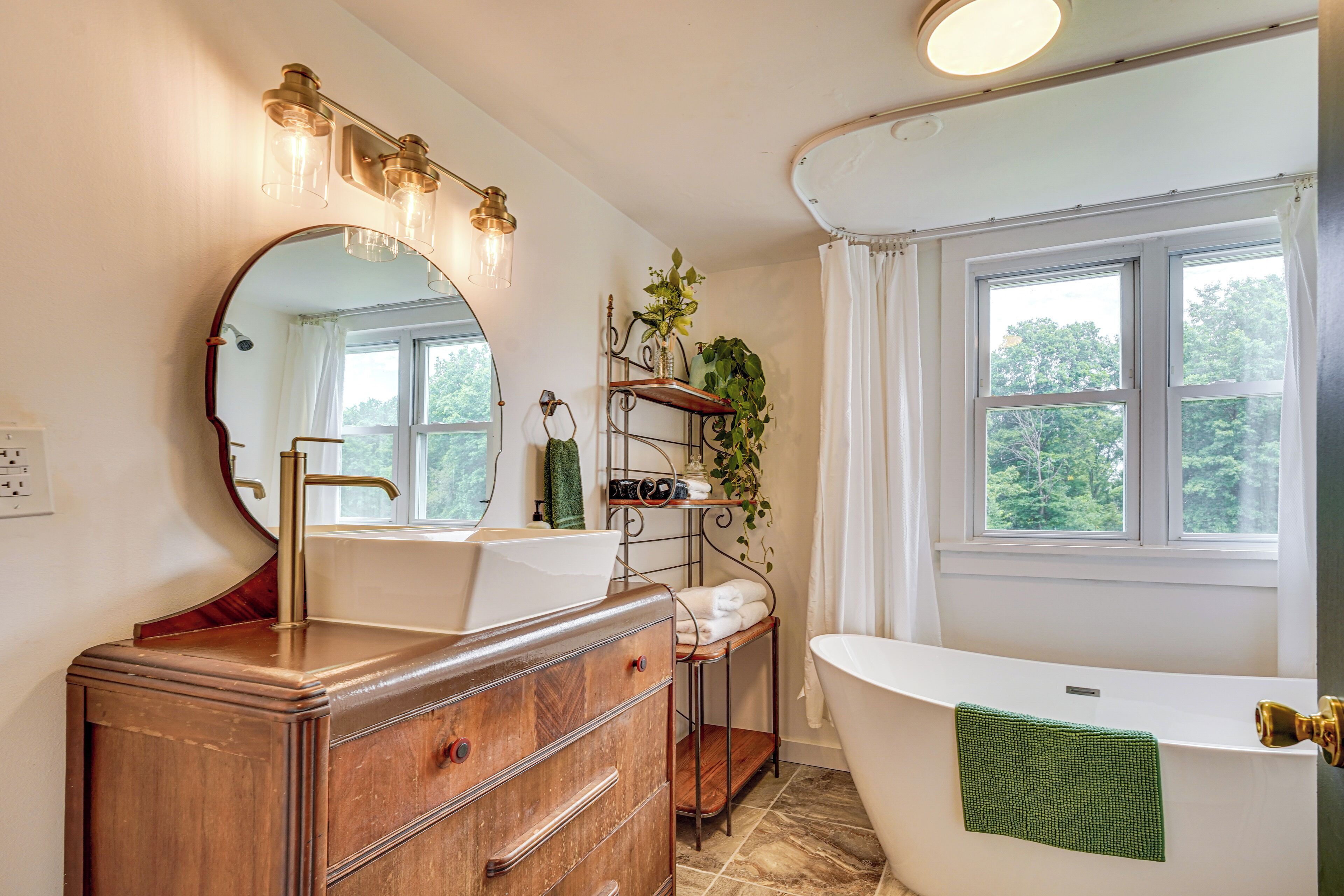 Un baño con un espejo grande, un tocador de madera, una bañera blanca y una ventana con cortinas.