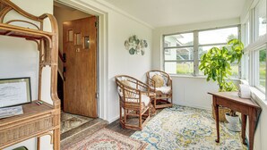 Une pièce avec une porte en bois, des chaises en osier, une petite table sur laquelle est posée une plante, et un tapis à motifs.