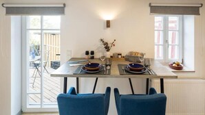 Comedor con una mesa dispuesta para dos personas, dos sillas azules, un florero con flores y una ventana con cortinas.