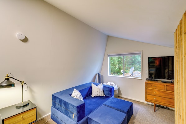 Quarto com sofá azul, cómoda de madeira, mesa de escritório com abajur e televisão.