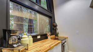 A kitchen with a wooden countertop, a coffee maker, a window with a view of trees, and a small shelf with bottles.