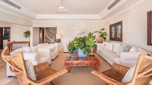 Un salón espacioso con un conjunto de sofá blanco, dos sillones de mimbre, una mesa de centro de madera y una planta grande.