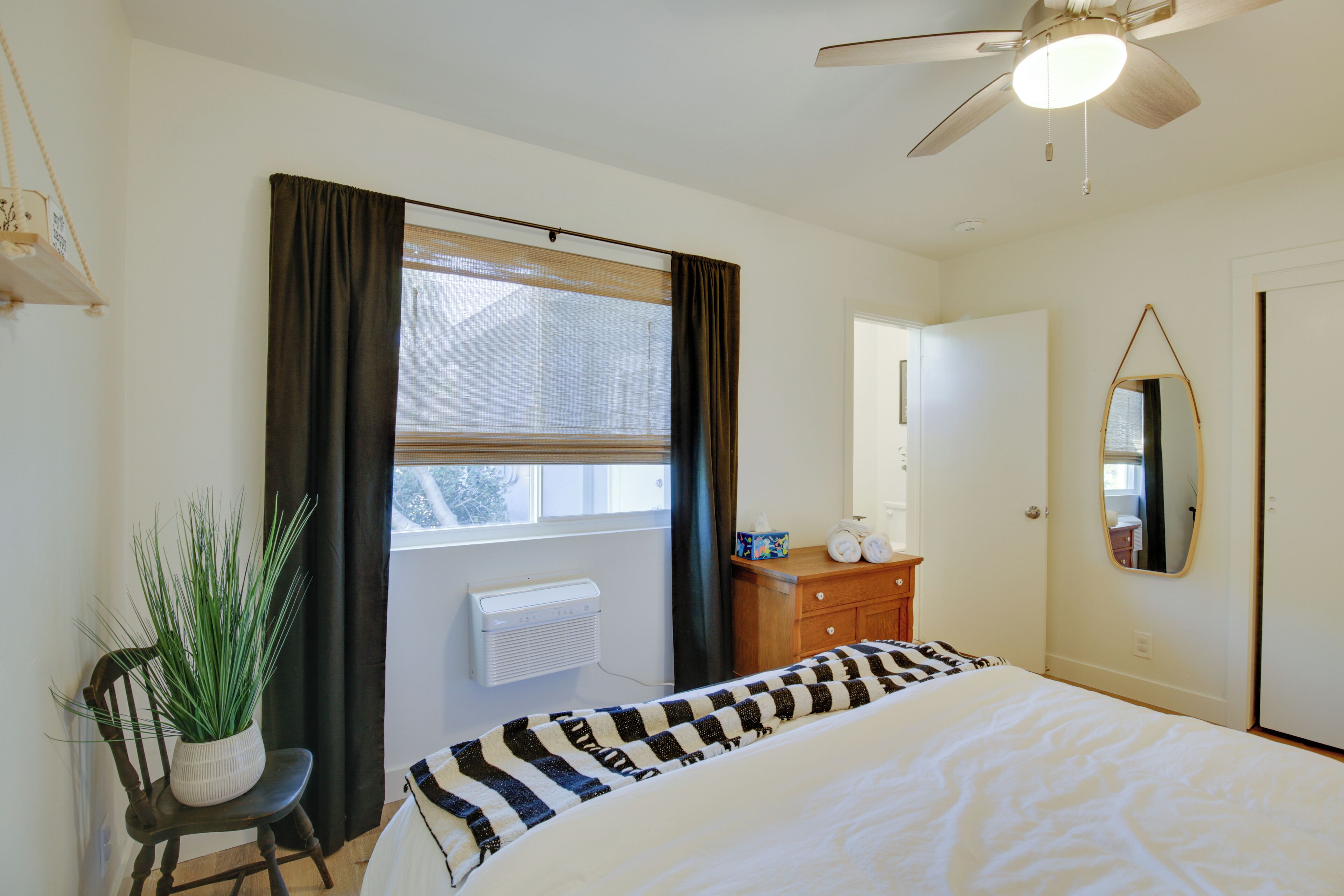 Une chambre à coucher comprenant un lit, un ventilateur de plafond, une fenêtre avec des rideaux, une commode, un miroir et une plante en pot.
