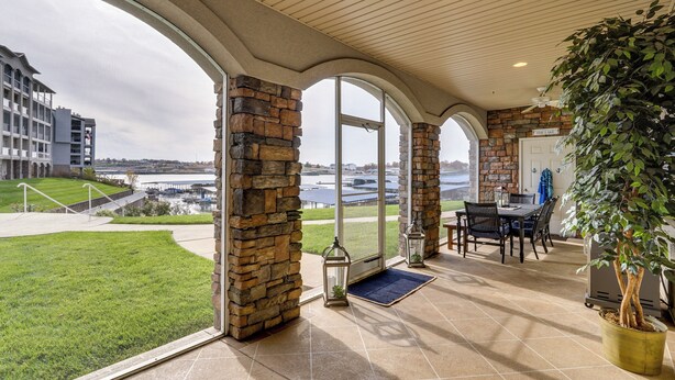 A covered patio with stone walls, large windows, a dining area, and a view of a body of water. - Boat Slip + Pool Access: Osage Beach Retreat (Osage Beach)