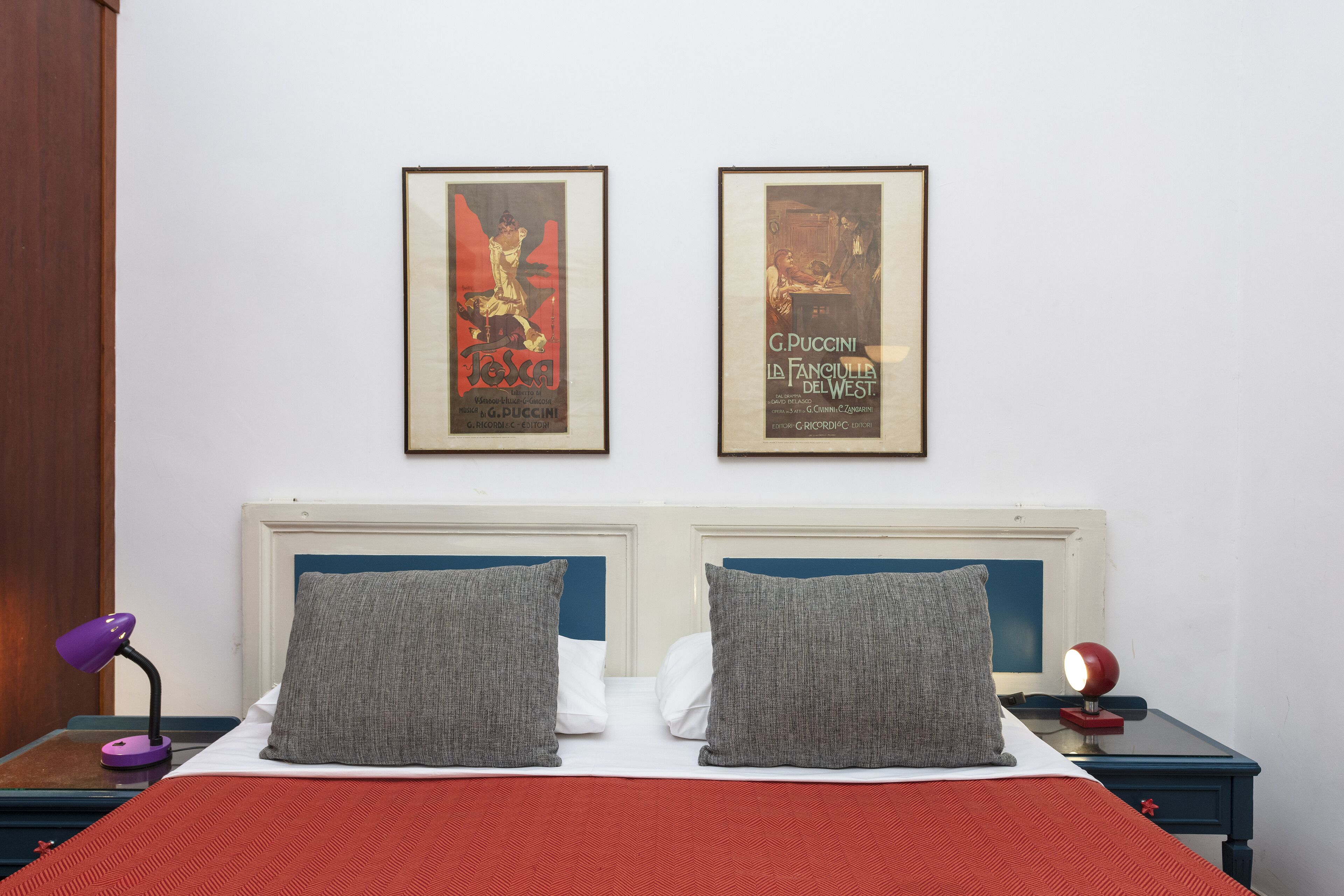 A neatly made bed with a red blanket, two grey pillows, and a white pillow. There are two framed posters on the wall above the bed.