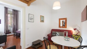 A cozy living area with a red sofa, a white table, and a wooden bench. There are framed pictures on the wall and a ceiling light. - Opera House (Rome)