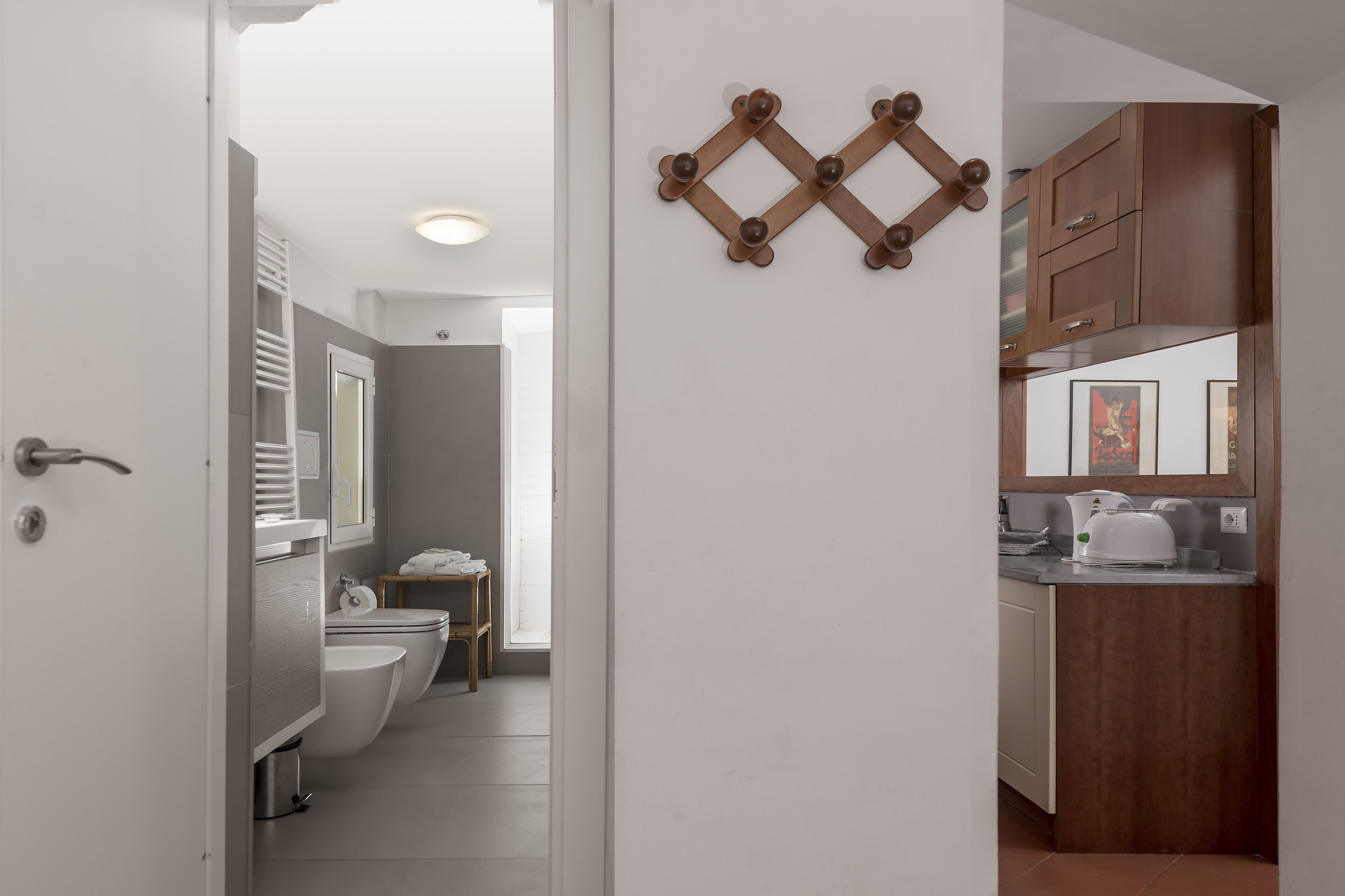 Een moderne badkamer met toilet, wastafel en spiegel. Er is een houten decoratie aan de muur en een kitchenette met kastjes en waterkoker.